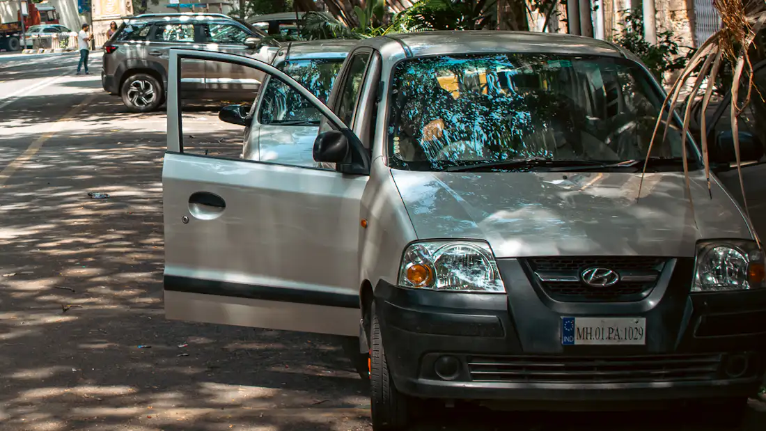 Old silver hatchback with a cracked windscreen parked on a tree-lined Mumbai street